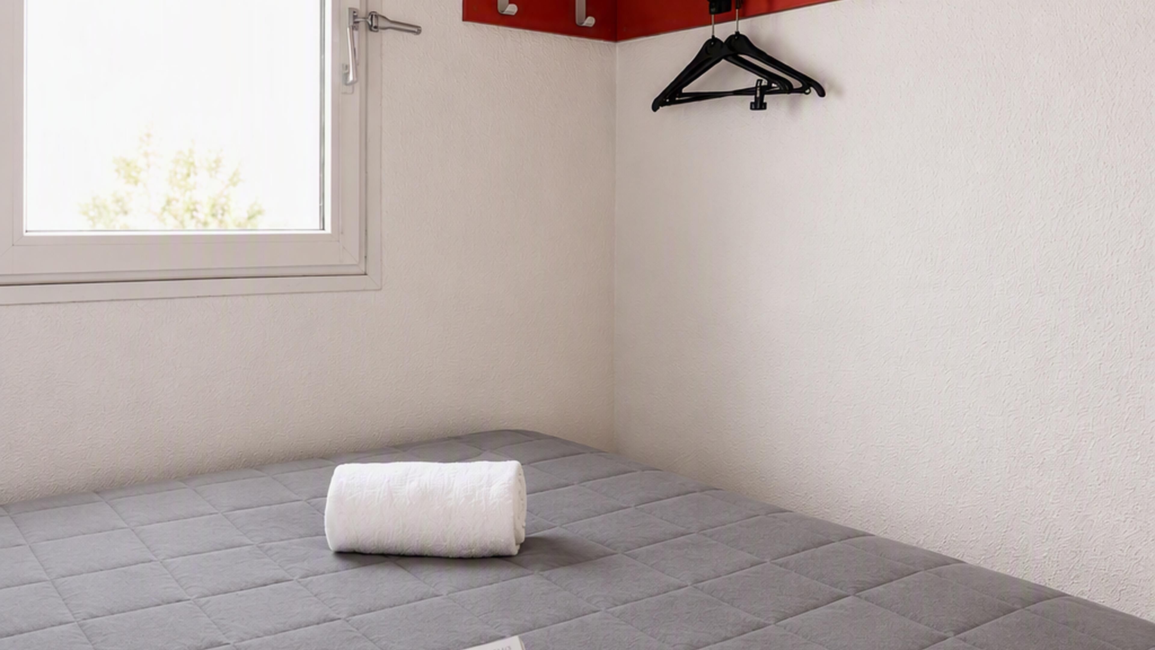 Una habitación pequeña con cama individual, colchón gris, toalla blanca y ventana con vista a árboles.