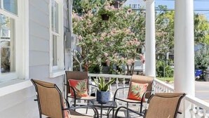 A covered porch with four chairs and a table, white railings, and a view of a tree with pink flowers. - Six on Cannon | 6BR Charleston Home + Parking (Charleston)