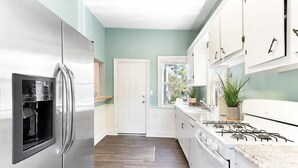 A kitchen with a stainless steel refrigerator, white cabinets, a gas stove, and a window with a view of greenery. - Six on Cannon | 6BR Charleston Home + Parking (Charleston)