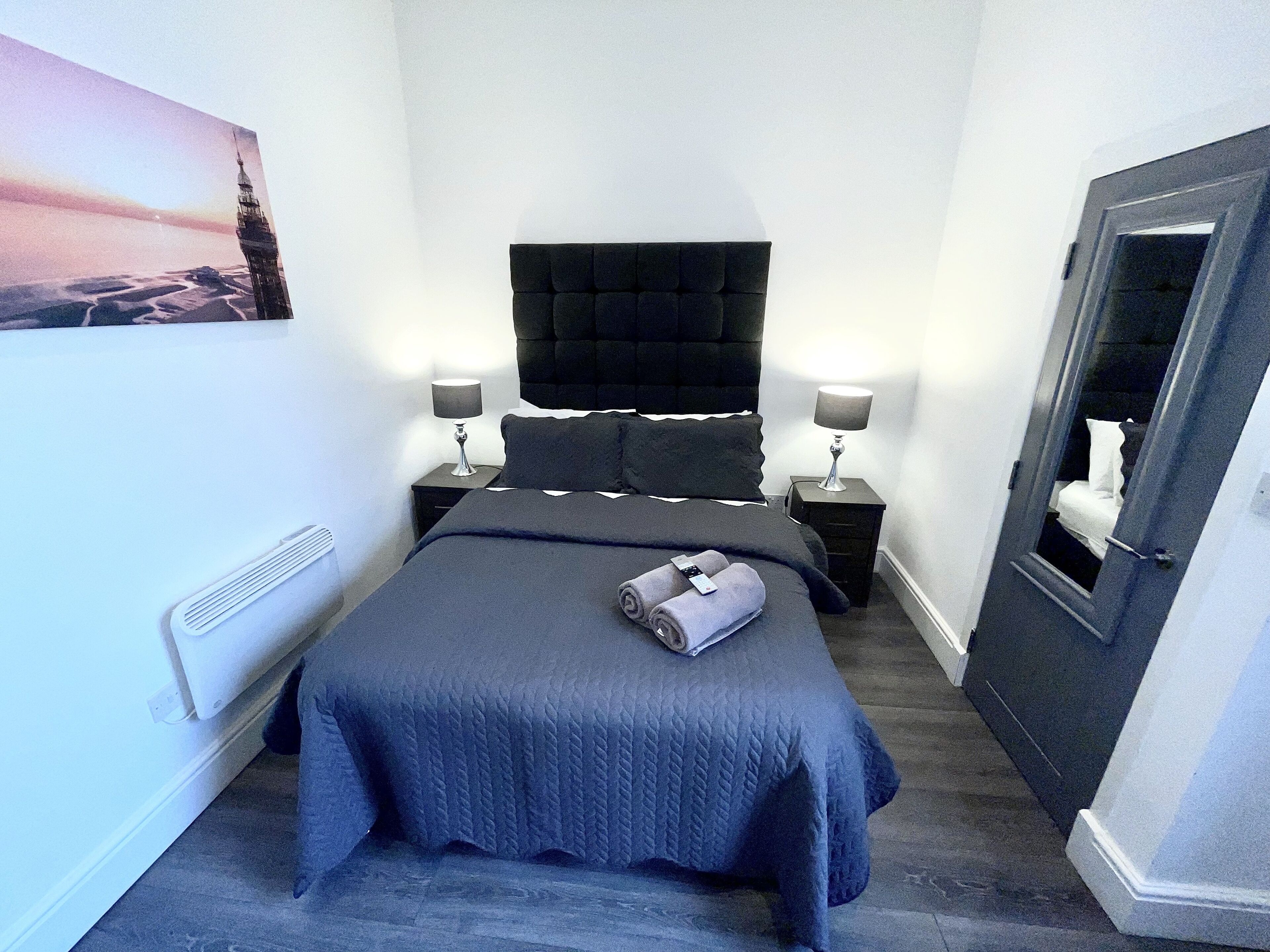 A neatly arranged bedroom with a large bed, two bedside tables with lamps, a dark headboard, a wall-mounted heater, and a door with a mirror.