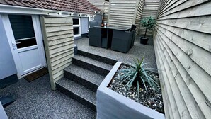 A compact outdoor patio with a small table and chairs, a concrete planter with a plant, and a set of steps leading to a door. - Albert Luxury Hottub and Jacuzzi Apartments (Blackpool)