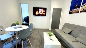 A modern living room with a grey sofa, a white coffee table with a plant, a round dining table with chairs, a TV mounted on the wall, and a door in the background. - Albert Luxury Hottub and Jacuzzi Apartments (Blackpool)
