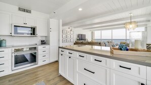 Een moderne keuken met witte kasten, een inbouwoven, een magnetron en een groot eiland met een spoelbak. Er is een zithoek met een bank en een groot raam met uitzicht op de oceaan.