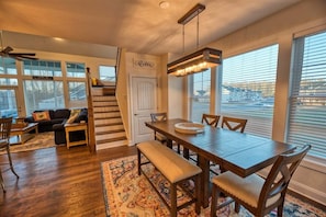 A spacious living area with a wooden dining table, benches, a staircase, and large windows.
