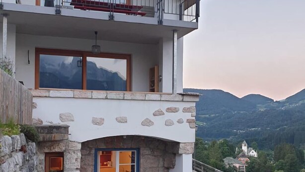 A two-story house with a stone facade, a balcony with a white umbrella, and a view of mountains in the background. - Ferienwohnung Oma Rosa mit Traumblick und Zentrumsnähe (Berchtesgaden)