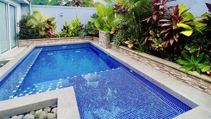 Una piscina rettangolare con piastrelle di mosaico blu, circondata da diverse piante tropicali e da un muro in pietra.