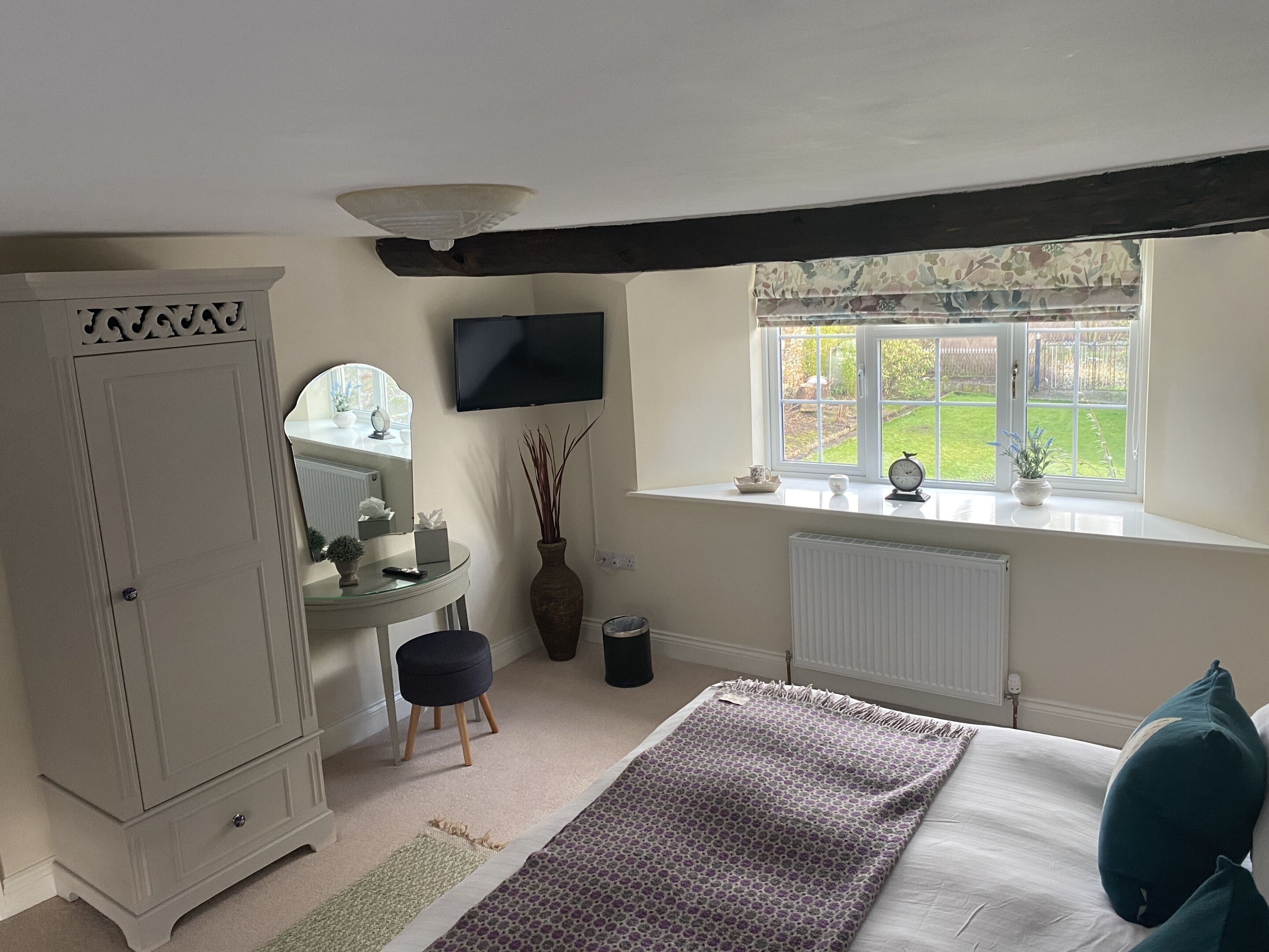 A bedroom with a bed, a dresser, a vanity with a mirror, a window with a view of the garden, and a television on the wall.