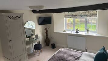 A bedroom with a bed, a dresser, a vanity with a mirror, a window with a view of the garden, and a television on the wall.