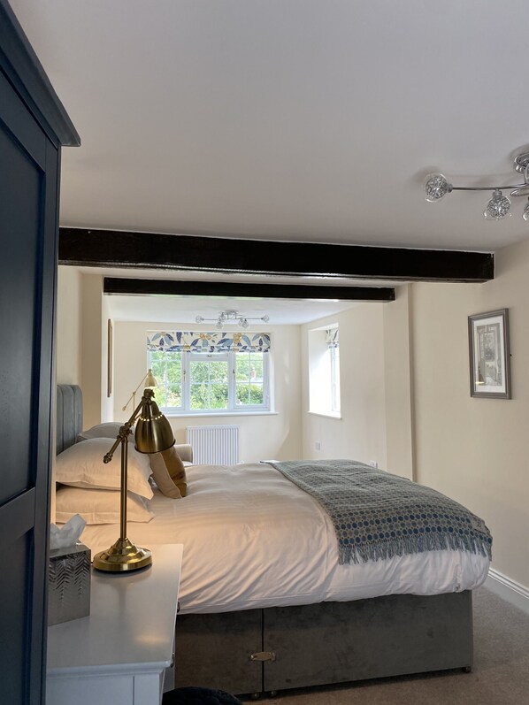 A bedroom with a bed, bedside lamp, and a window with a view of greenery. - Cosy Nook Cottage (Appleby-in-Westmorland)