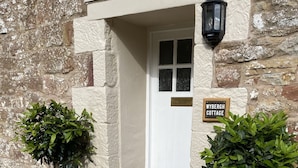 Un bâtiment en pierre avec une porte blanche, deux plantes en pot et une enseigne indiquant « Wybergh Cottage ».