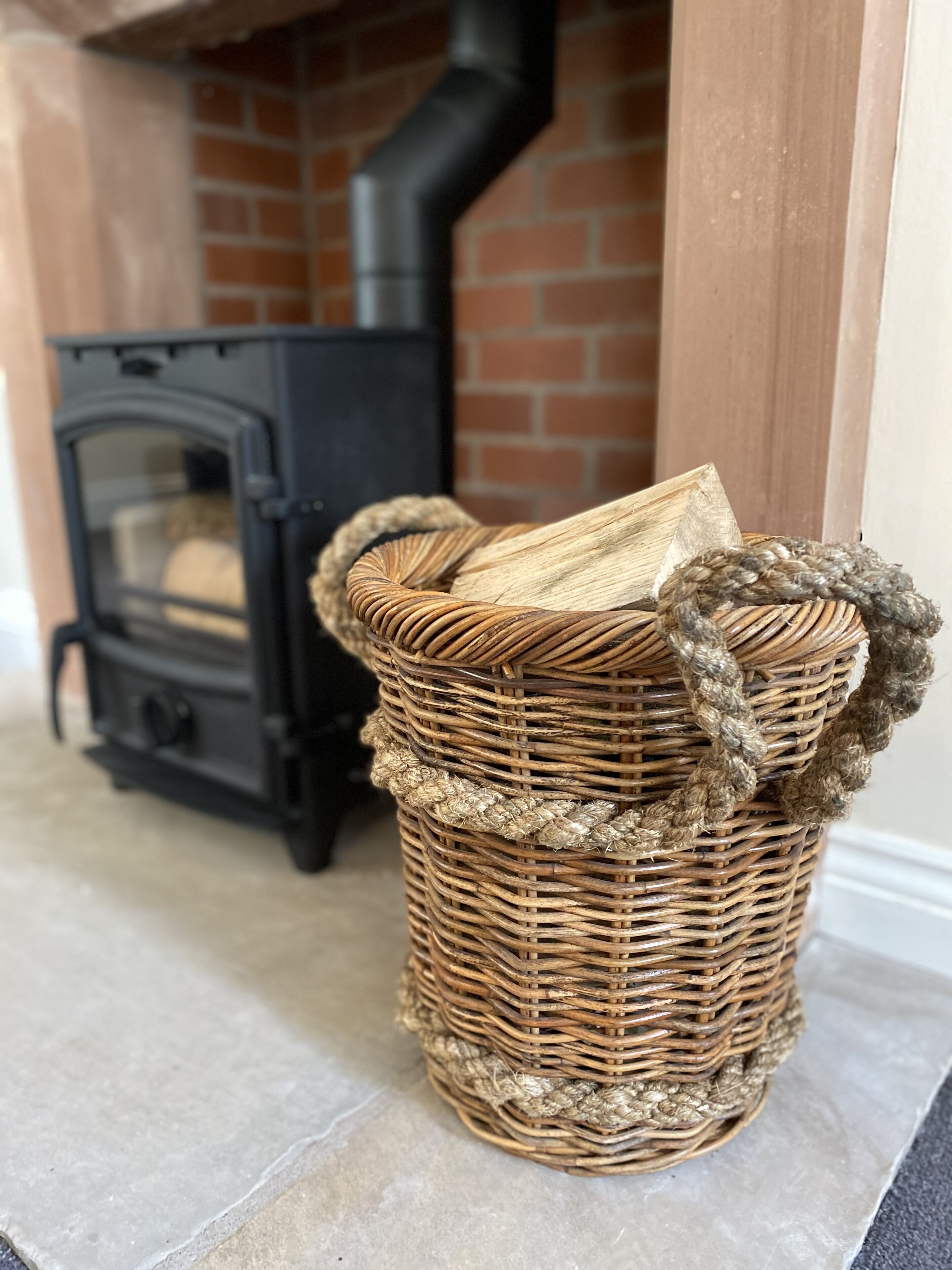 A wicker basket with a rope handle on a tiled floor, a black wood-burning stove, and a brick wall in the background.