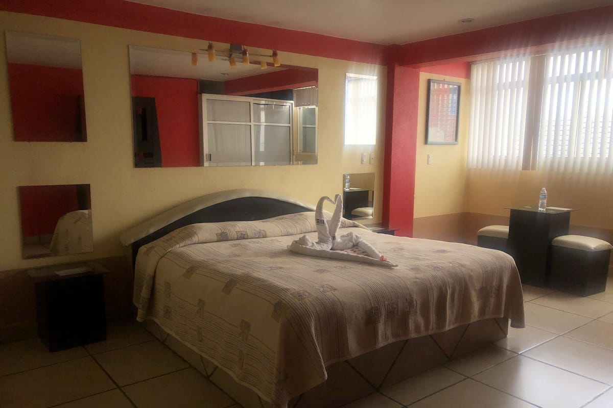 A hotel room with a bed, a bedside table, a mirror, a window with curtains, and a small table with a bottle.