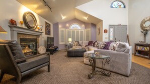A spacious living room with a fireplace, comfortable seating, a glass coffee table, and a large window with shutters. - Harvest House Inn (Springdale)