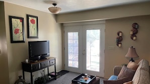 A living room with a television on a stand, a coffee table, a sofa, and a door leading to a balcony. - Harvest House Inn (Springdale)