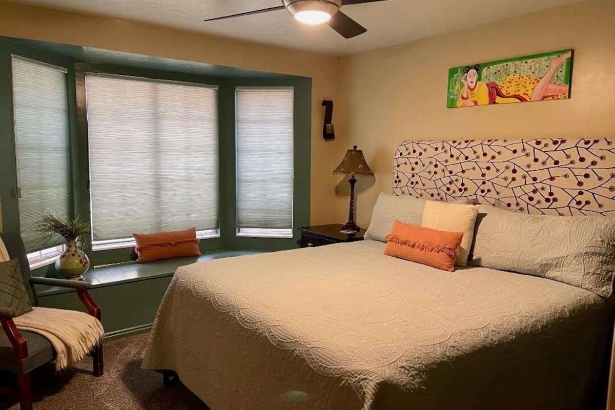 A bedroom with a bed, a chair, a window with blinds, a ceiling fan, and a wall with a patterned design.