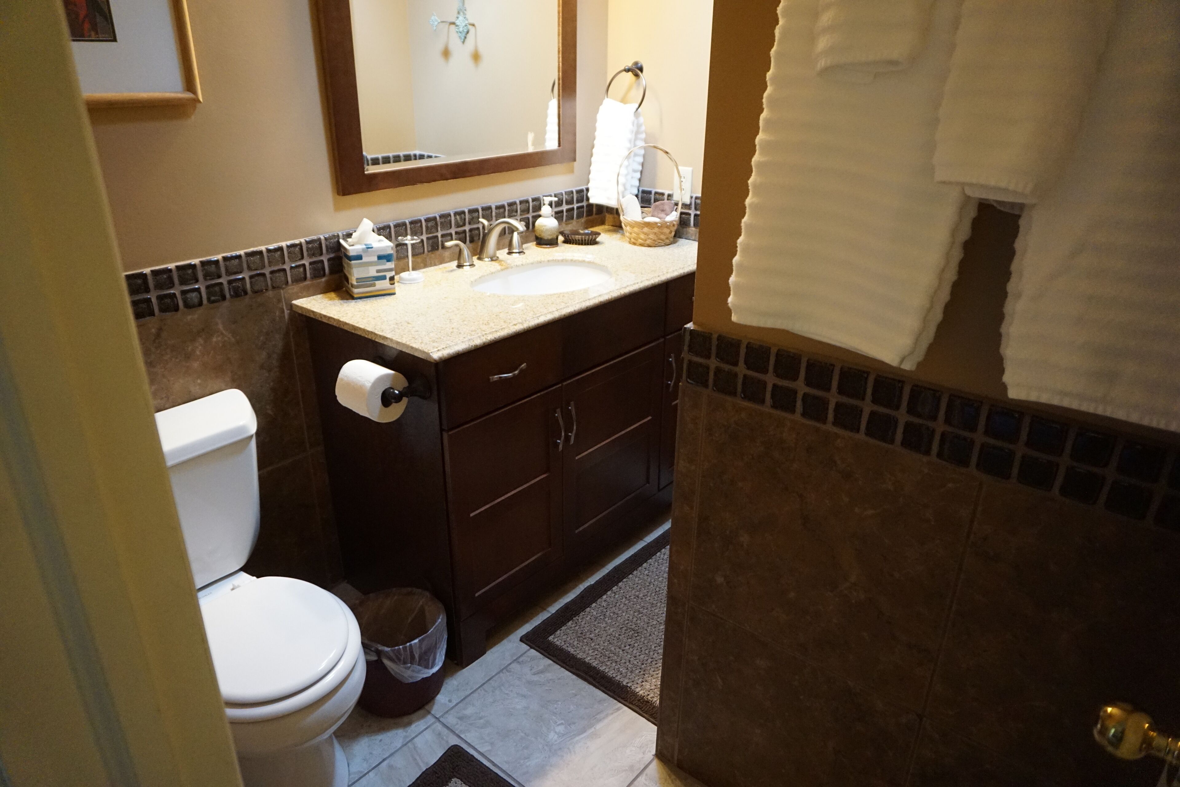 A bathroom with a toilet, sink, and mirror. There are towels hanging on the wall and a trash can.