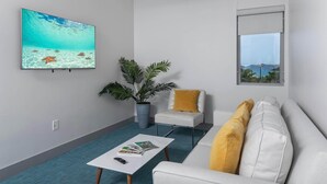 A modern living room with a large wall-mounted TV, a white sofa with yellow cushions, a small white chair, a coffee table with magazines, and a large potted plant. - 2 Bedroom, Sleeps 6 Guests (Cruz Bay)