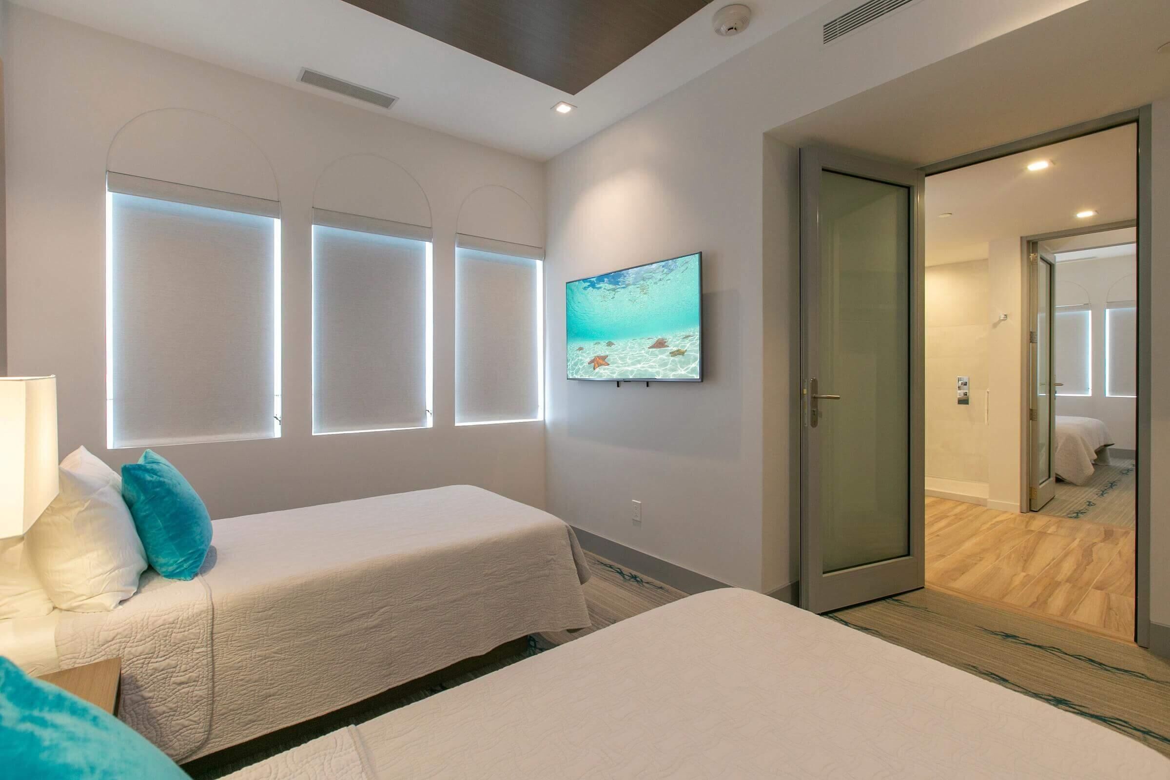 A hotel room with two beds, a large window with a view, a painting on the wall, and a door leading to a bathroom.