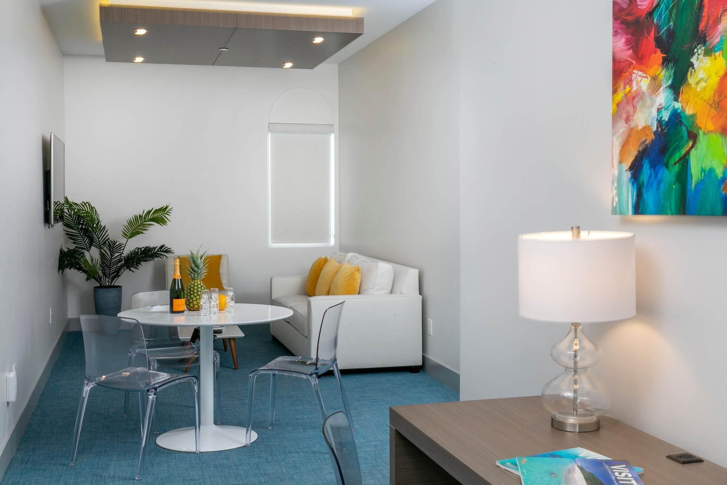Una camera d'albergo moderna con un divano bianco, un tavolo da pranzo con sedie, una pianta in vaso, un quadro astratto dai colori vivaci e una lampada.