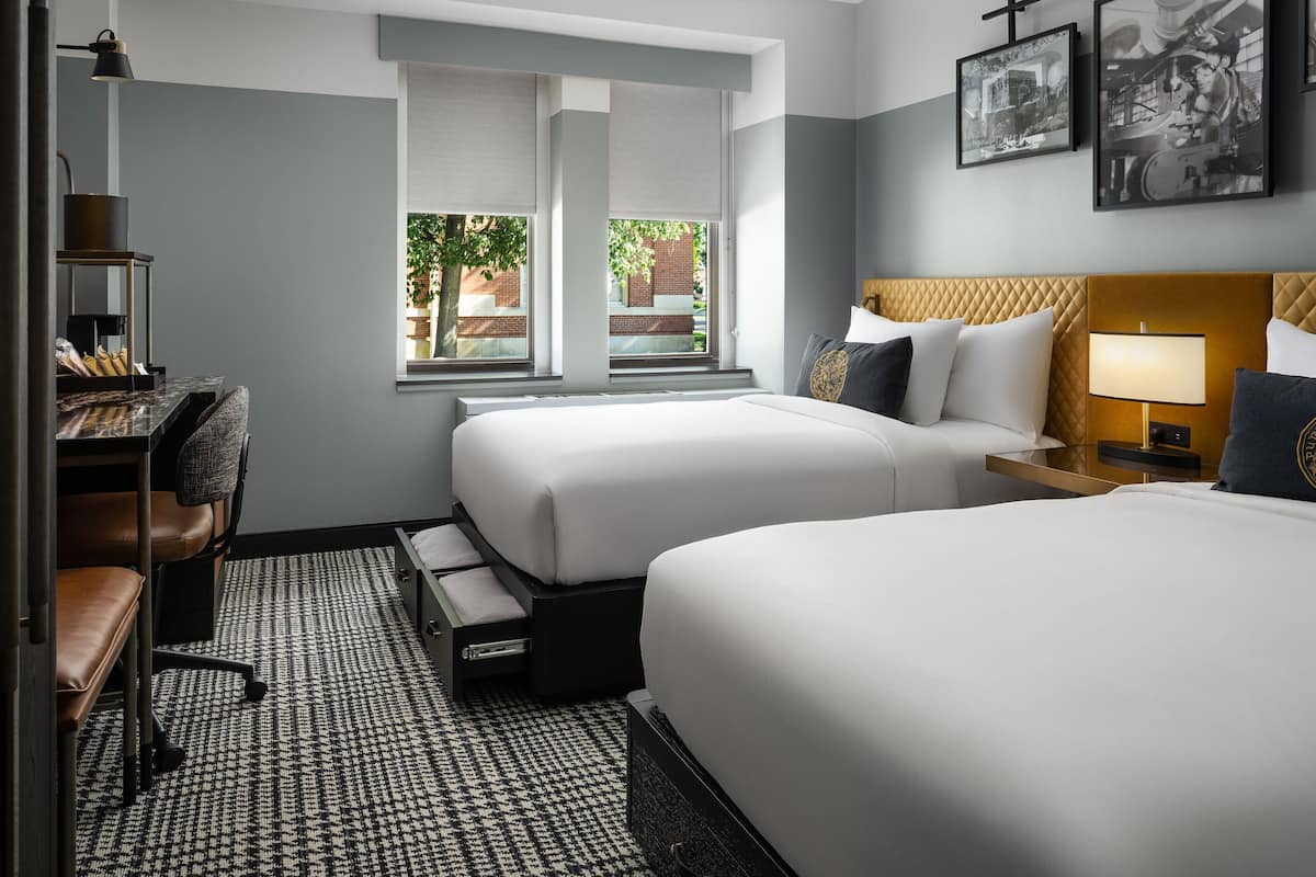 Habitación de hotel con dos camas, un escritorio, una silla, una lámpara y una ventana con vista a áreas verdes.