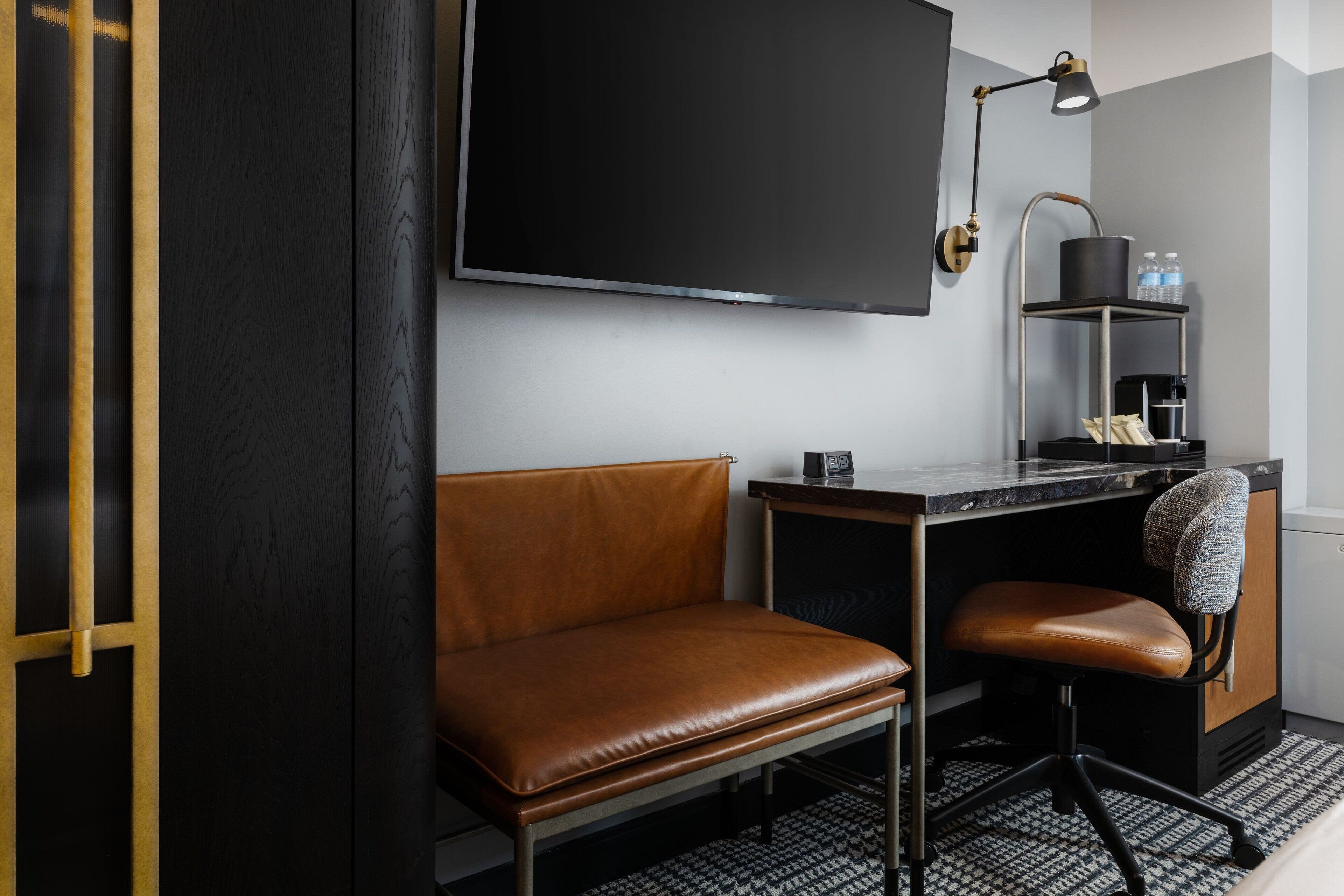 Ein modernes Zimmer mit einer Ledersofa, einem Schreibtisch mit Computer, einem Wand-Fernseher und einem kleinen Regal mit Gegenständen.