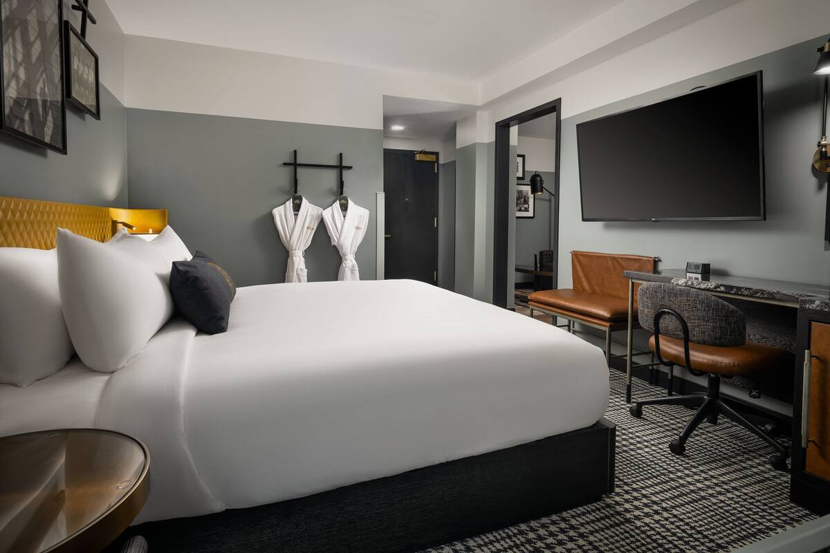 A hotel room with a large bed, a desk with a chair, a TV, and two robes hanging on hooks.