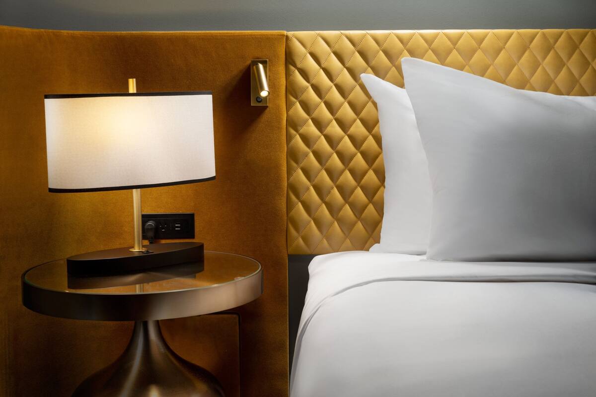 A hotel room with a round bedside table, a lamp, a bed with white bedding, and a quilted headboard.