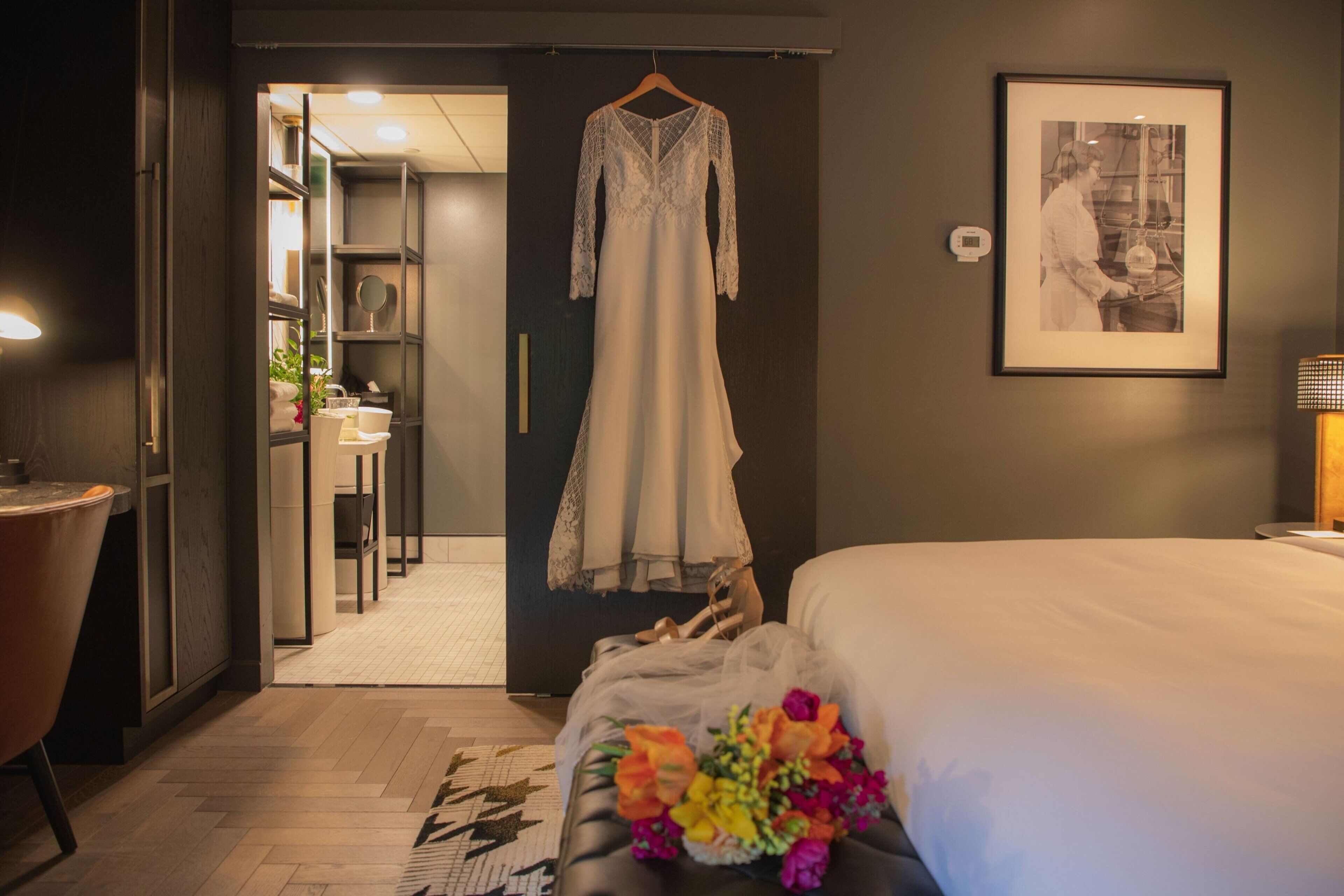 A hotel room with a bed, a hanging dress, a framed picture, and a desk with a lamp.