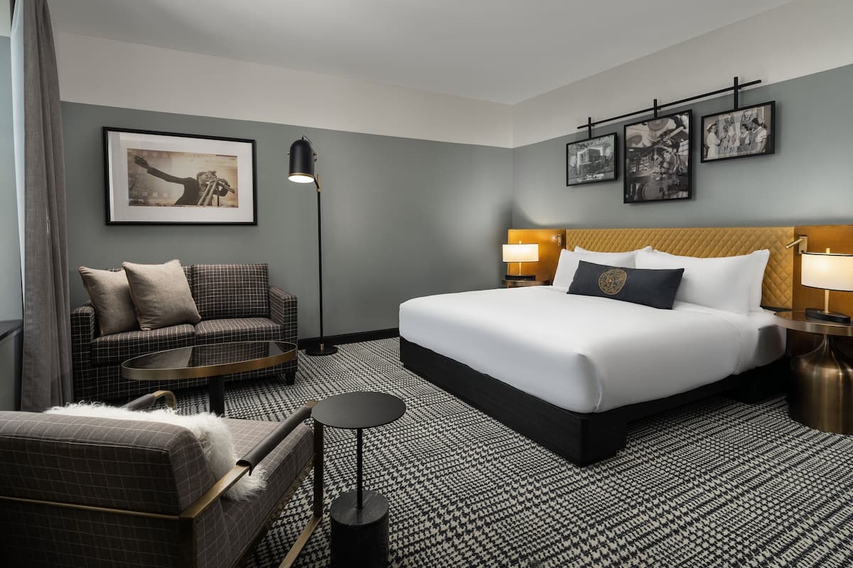 Habitación de hotel con una cama grande, un sofá, una mesita y una lámpara. En la pared hay cuadros enmarcados.