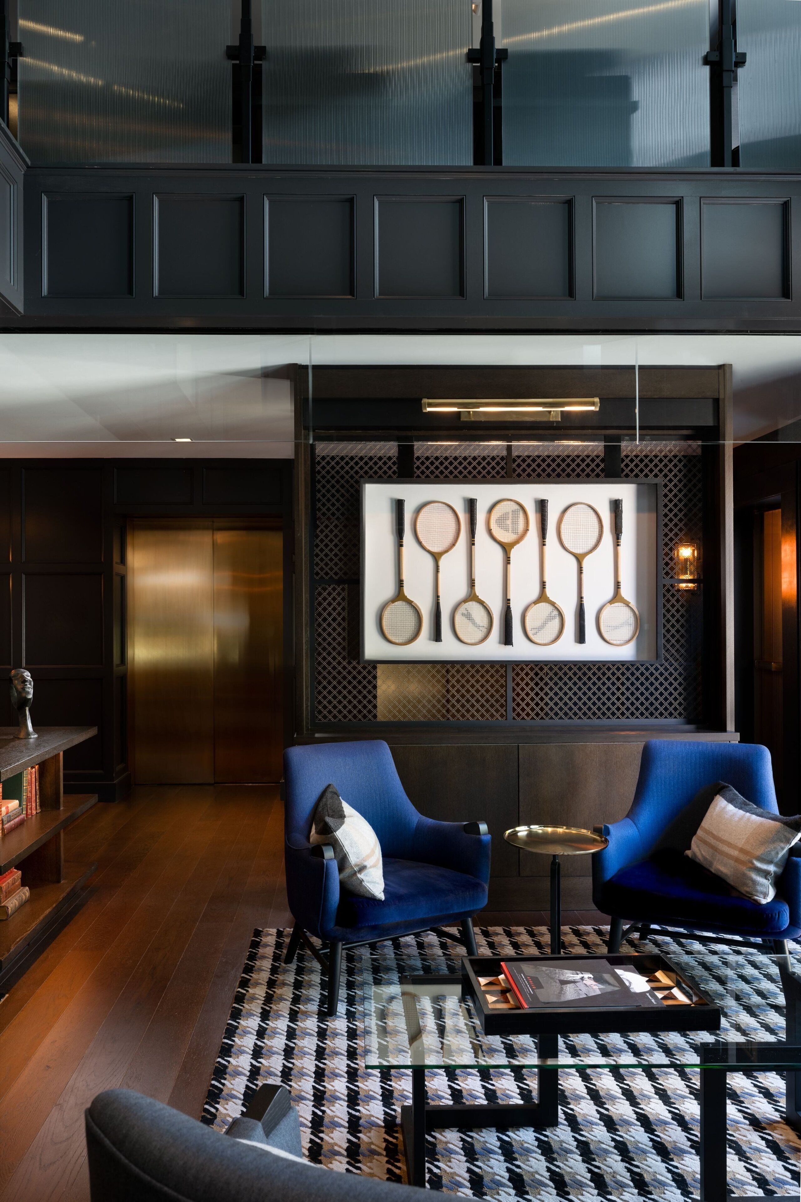 A modern lobby with two blue armchairs, a patterned rug, a glass coffee table, and a wall-mounted tennis racket display.