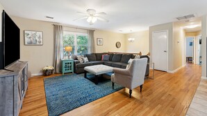 A spacious living room with a sectional sofa, ottoman, and a television on a wooden cabinet. - Two Tides | Two Units in One | Screened In Porch | Steps to the Beach! (Folly Beach)