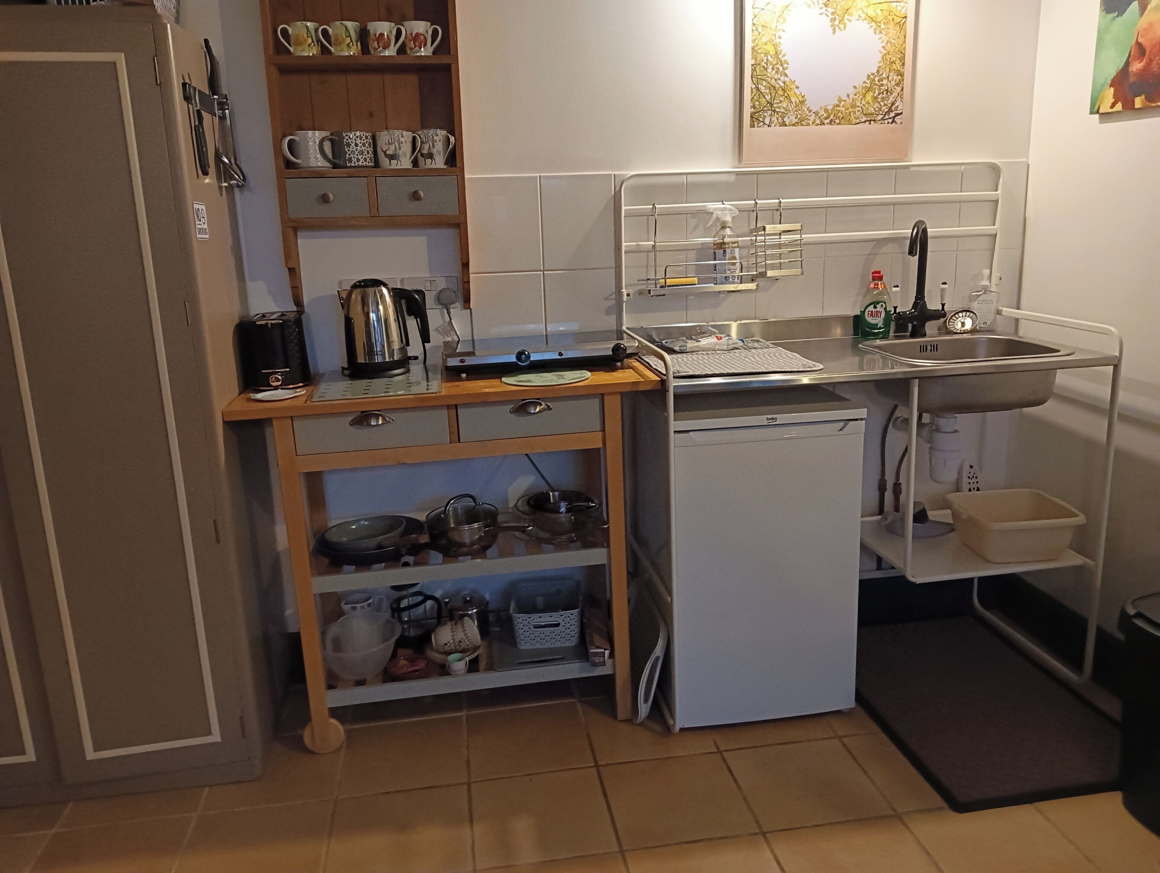 A compact kitchen with a dishwasher, a sink, and a small countertop. There are shelves with kitchenware and a kettle.
