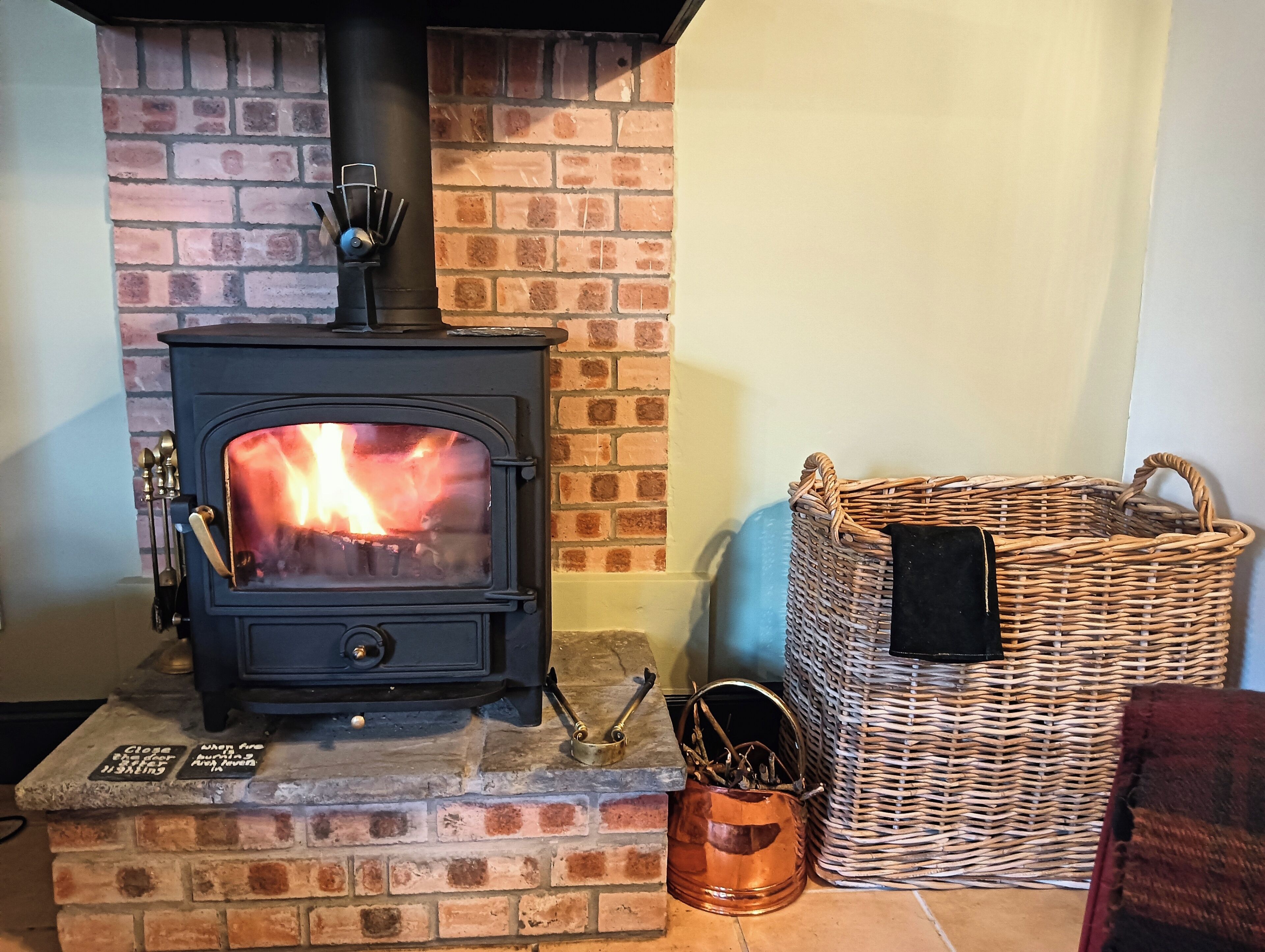 A wood-burning stove with a glass door, a copper pot, and a wicker basket.