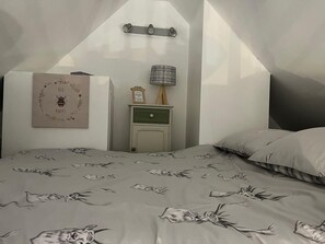 Un dormitorio con techo inclinado, una cama con cubrecama estampado con motivos florales, una mesita de noche con lámpara y una estantería en la pared con un cuadro enmarcado.