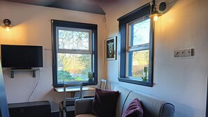 A living room with a grey sofa, a TV on a stand, a desk with a chair, and two windows with curtains. - Claymires Bothy Cottage Nr Loch Lomond & Stirling (Stirling)