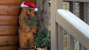 Une statue d’ours en bois portant un bonnet de Père Noël et ornée d’une couronne, un petit sapin de Noël et un mur en pierre.