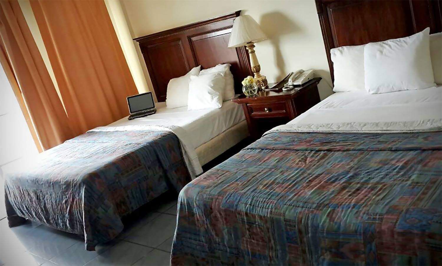 A hotel room with two beds, a nightstand with a lamp, a phone, and a laptop on one bed.