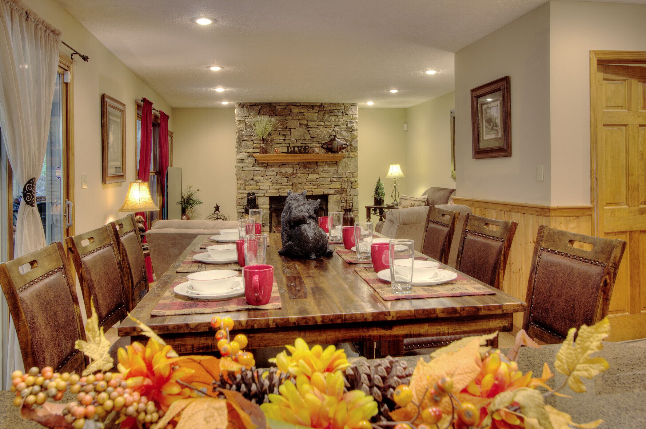 A dining room with a wooden table set for four, surrounded by chairs. A stone fireplace with a 'LIVE' sign above it is in the center.
