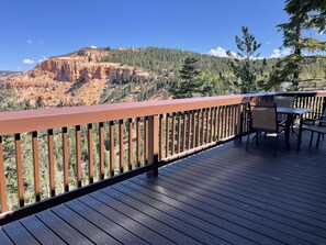 Una terraza de madera con barandilla, mesa y sillas, con vistas a un paisaje montañoso con árboles y cielo despejado.