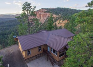 Une cabane à deux étages, avec des fondations en pierre, une façade marron et un toit en métal. Elle est entourée d’une forêt dense et offre une vue sur une vallée dominée par une falaise imposante.