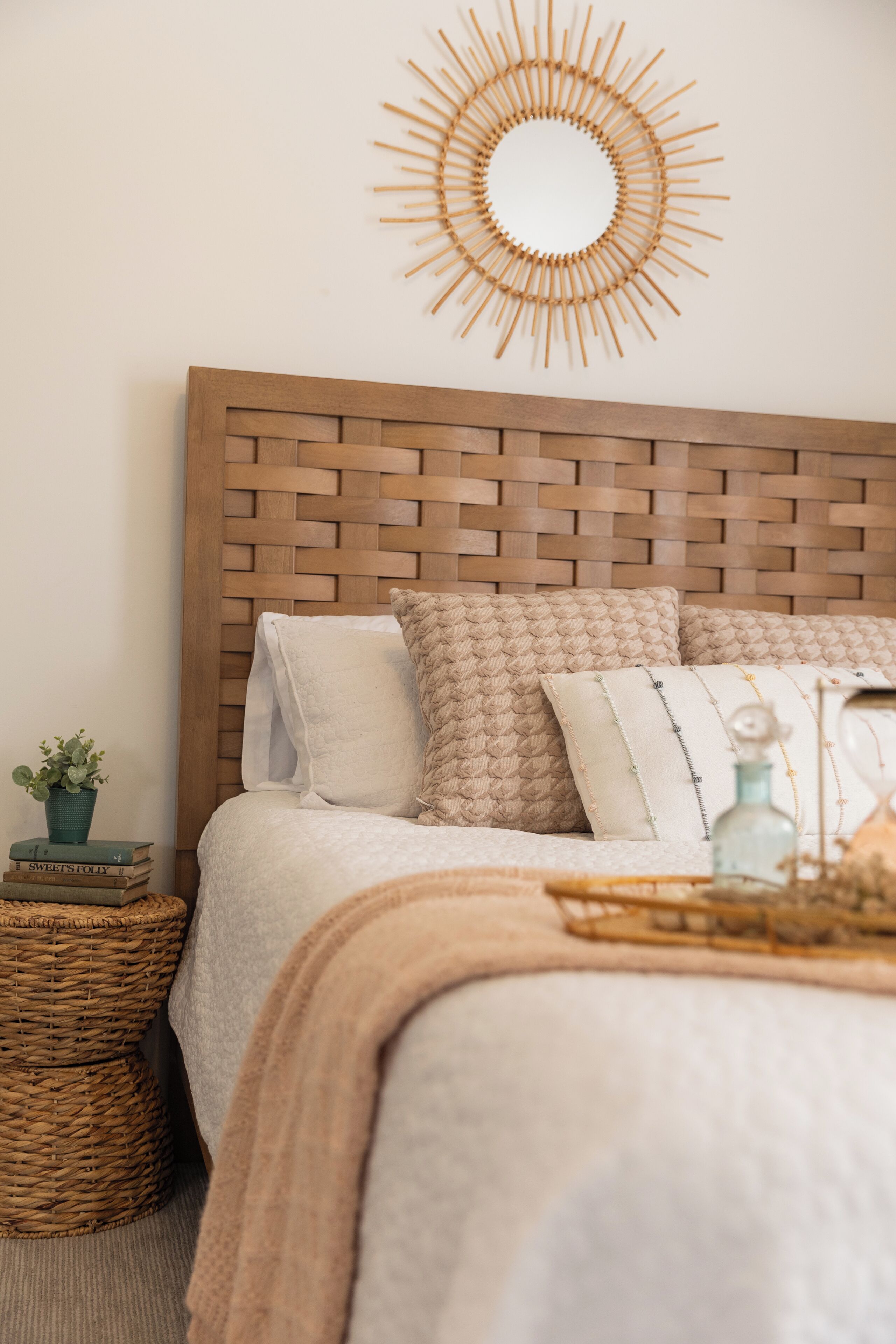 A neatly made bed with a woven headboard, a decorative wall piece, and a small side table with a plant and books.