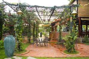 An outdoor patio area with brick flooring, tables, chairs, and a pergola covered with climbing plants.