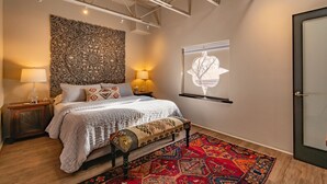 Een slaapkamer met een bed, nachtlampjes, een decoratief wandtapijt, een kunstwerk aan de muur, een bankje en een kamerbreed tapijt met patroon.