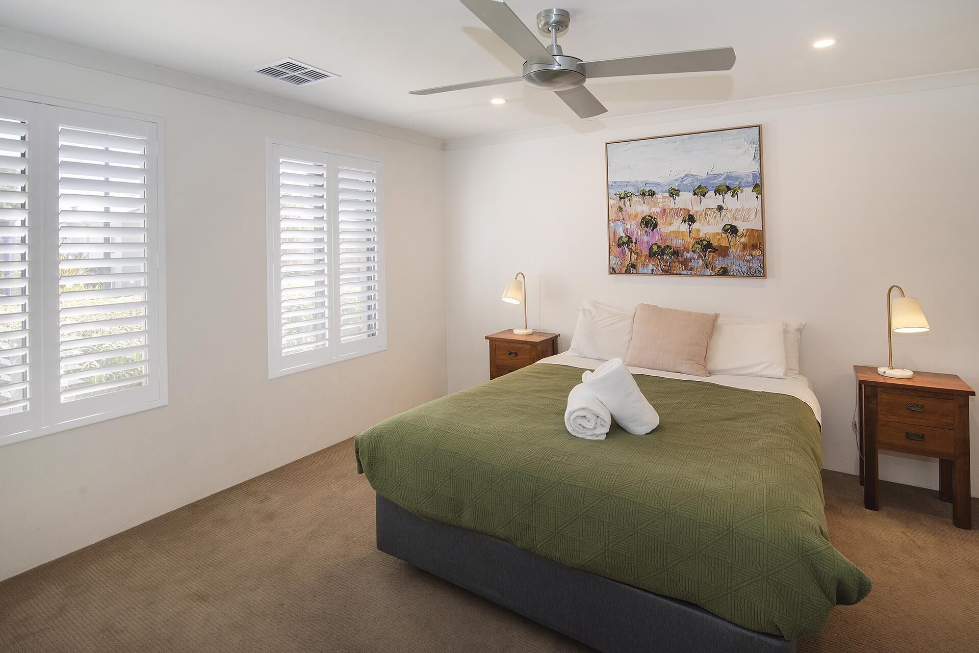 Une chambre à coucher avec un lit, deux tables de chevet, un ventilateur de plafond et un tableau accroché au mur.