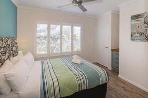 Ein Schlafzimmer mit einem Bett, einem Deckenventilator, einem Fenster mit Jalousien, einer Tür und einem kleinen Schrank.