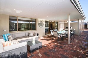 Ein überdachter Patio-Bereich mit Esstisch und Stühlen, einer Couch mit Kissen sowie einem großen Fenster.