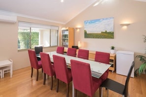 A dining area with a long table, red upholstered chairs, a white tablecloth, a small white refrigerator, and a window with a view of greenery. - House 20 - The Garden House Dunsborough (Dunsborough)