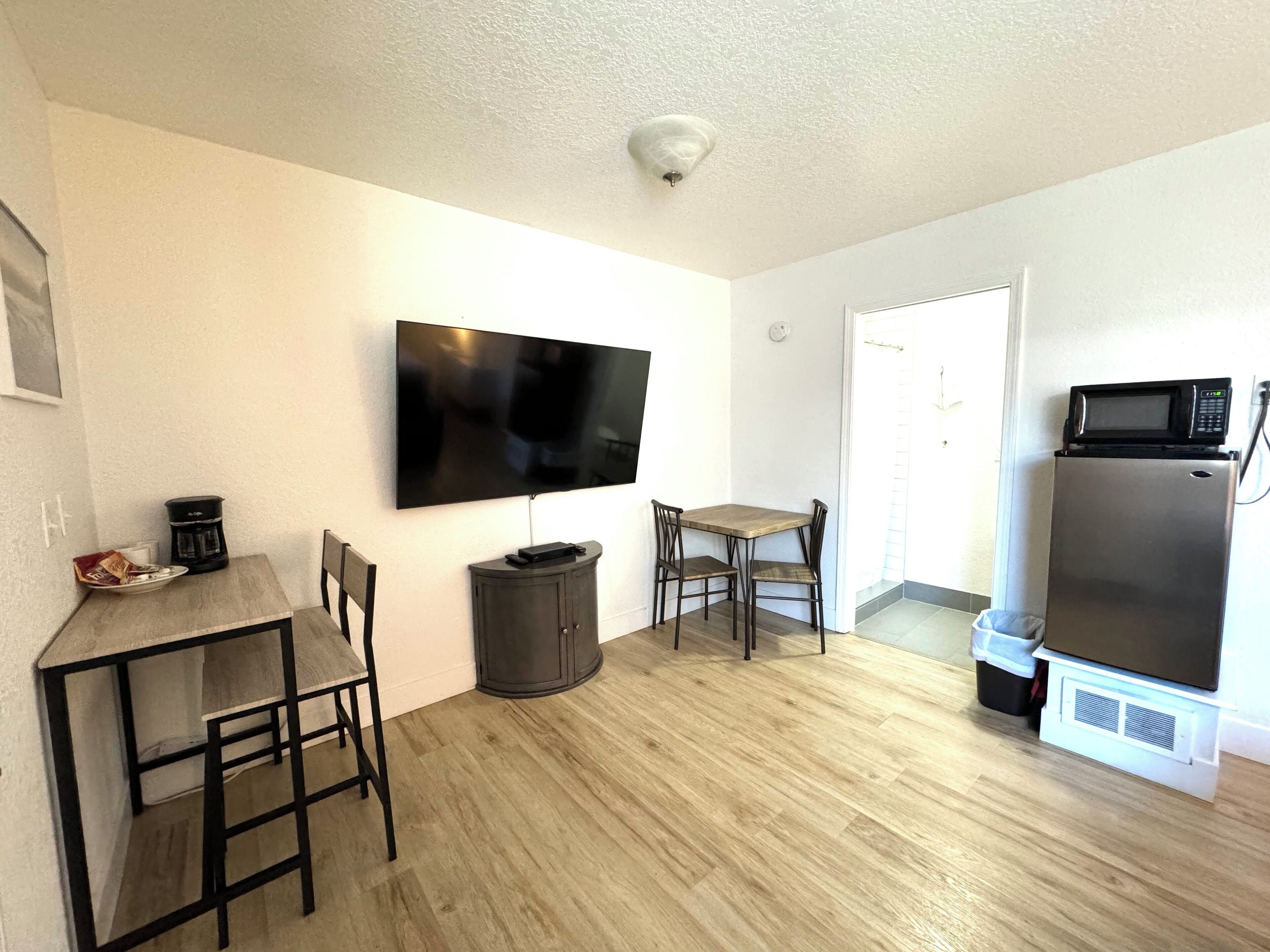 A compact kitchen with a flat-screen TV, a microwave, and a refrigerator. There is a dining table with chairs and a coffee maker.