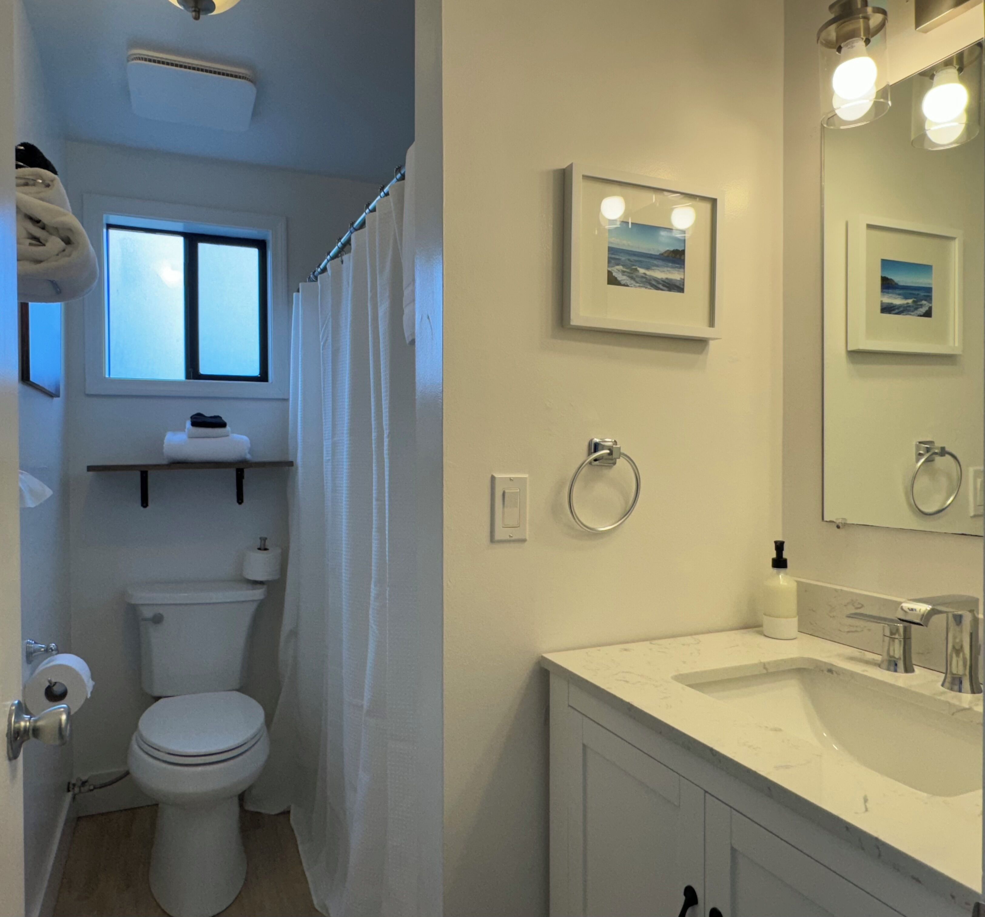 A bathroom with a toilet, sink, and shower curtain. There are framed pictures on the wall and a window above the toilet.