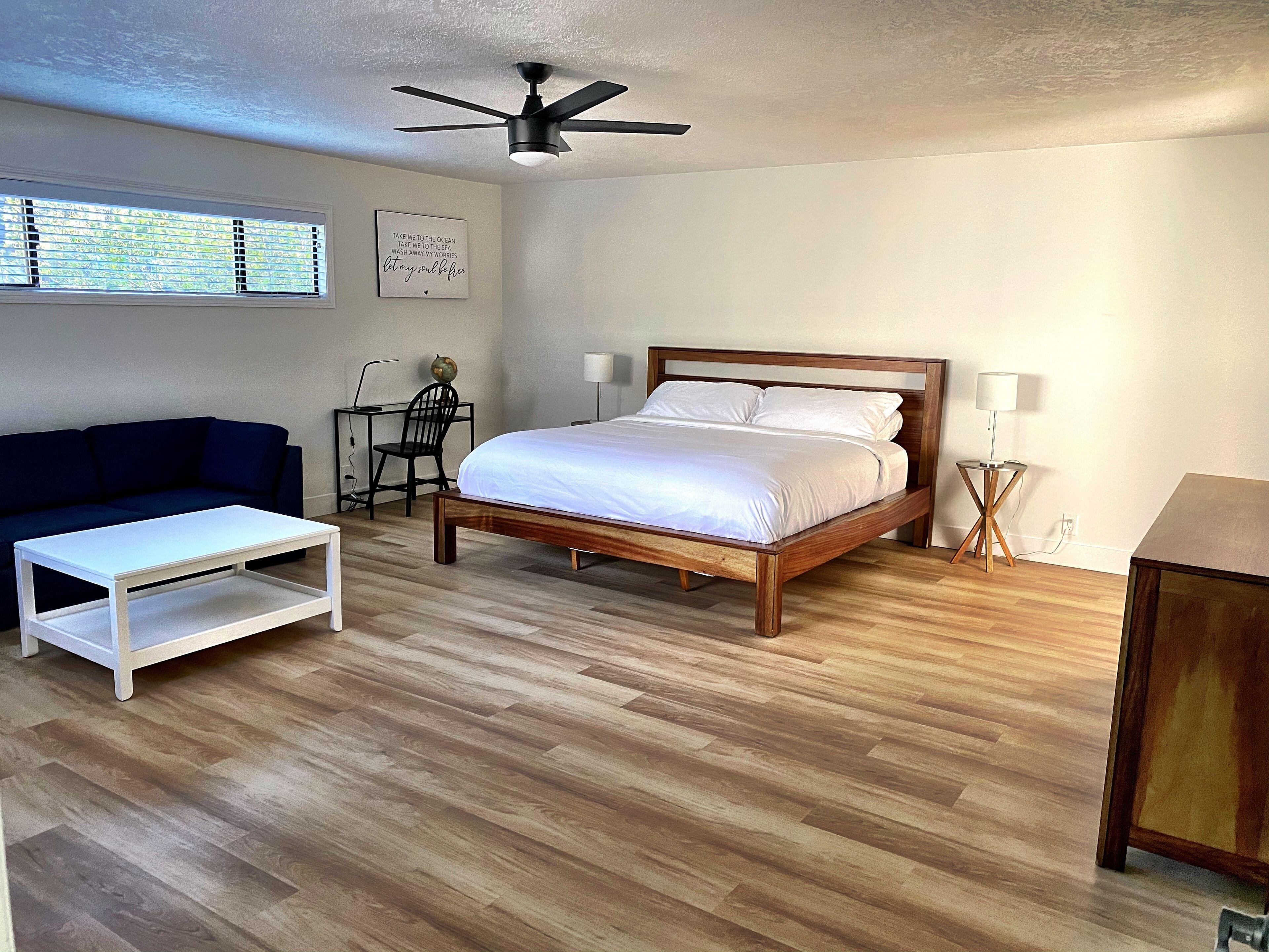 A bedroom with a wooden bed, a white sofa, a small table, a desk with a chair, and a ceiling fan.